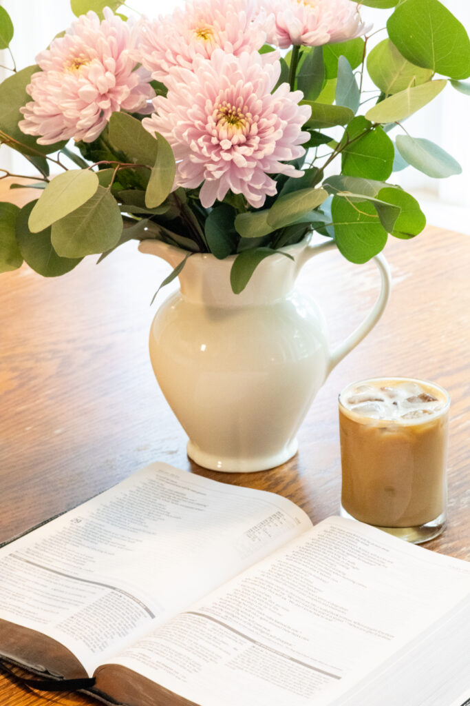 An open bible on the table with an iced coffee and white vase with flowers